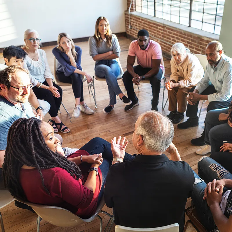 Diverse group of people sitting in a circle in a bright room discussing mental health support services.