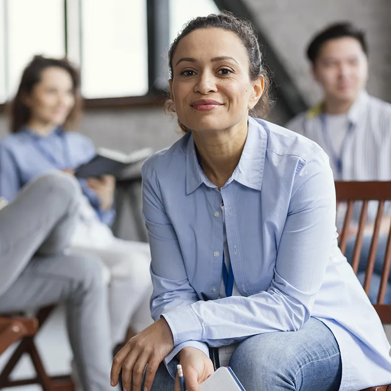 Close up of a woman in a light power blue blouse and blue jeans. In the background are other people in soft focus.