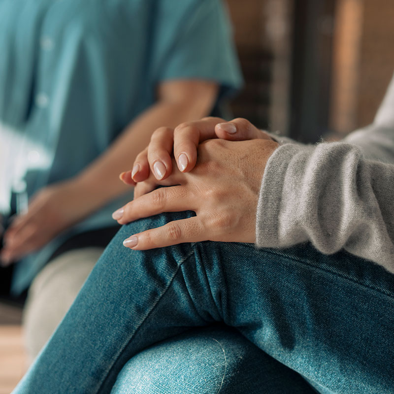 A woman, seated and dressed in blue jeans and a grey sweater, places both her hands on her knees.